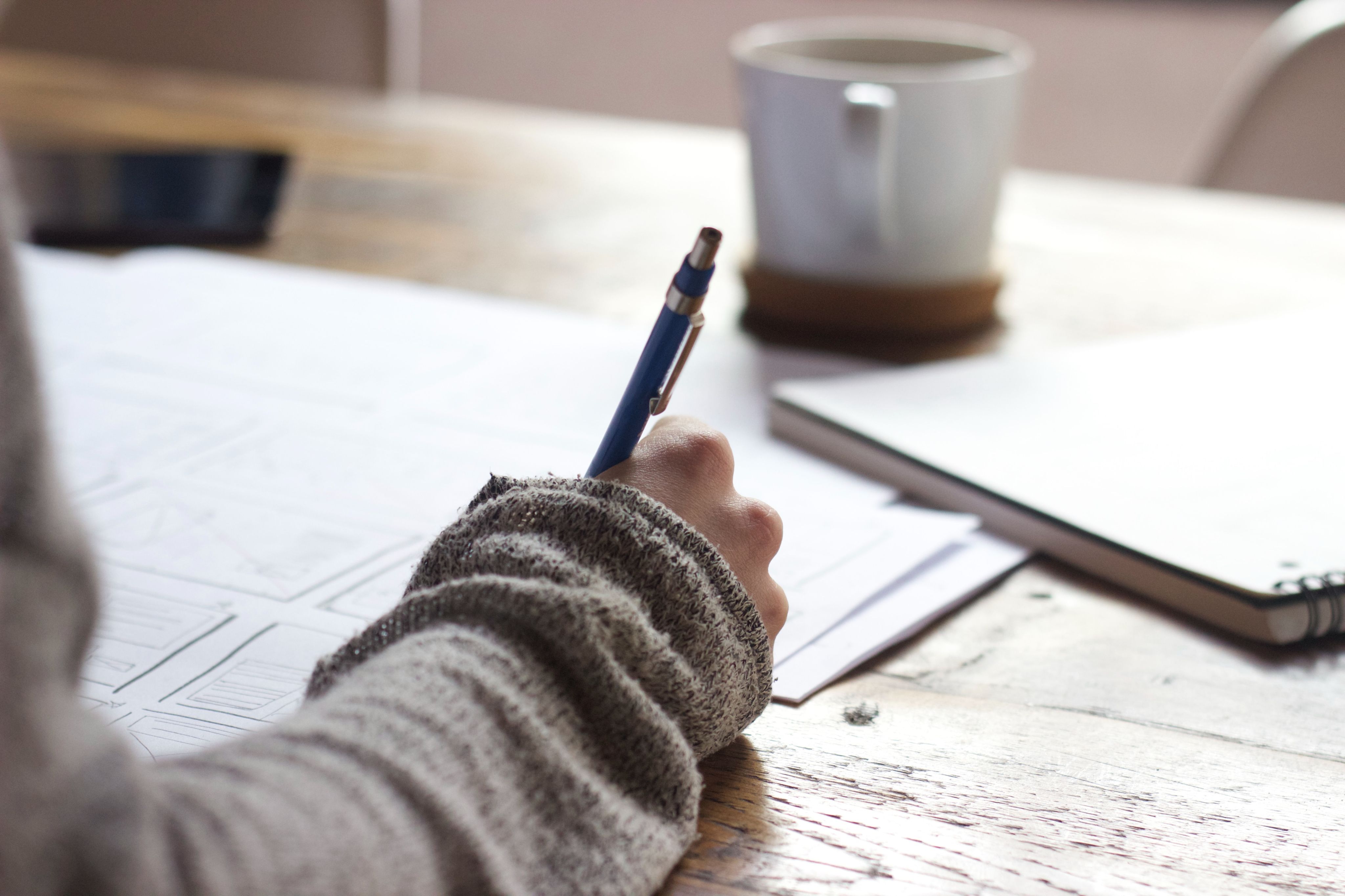 A hand holding a pen makes notes with a mug on a desk in the background