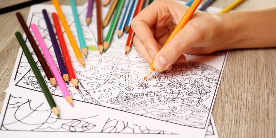Mindful colouring - a hand holds an orange colouring pencil and colours in a flower on a sheet of white paper with other colouring pencils laid beside