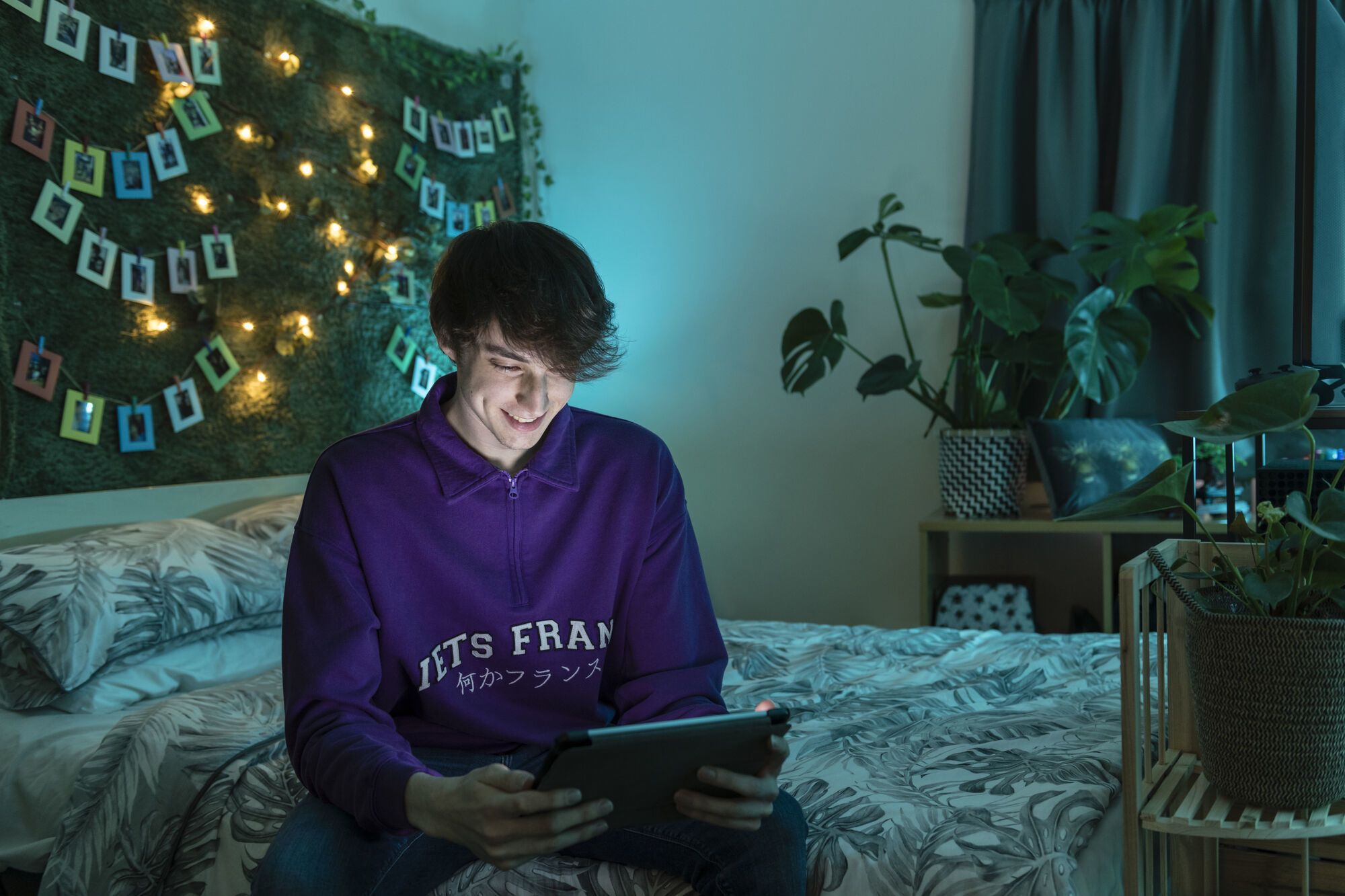 A male student looks at a tablet while sitting on a bed in a campus bedroom